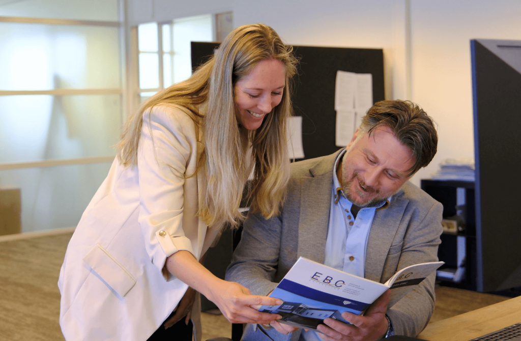 Beau, verpakkingsspecialist bij EBC Golfkarton, aan het werk tijdens een klantbezoek; hij bespreekt een maatwerk doosontwerp met een klant in een magazijn vol verpakkingen.