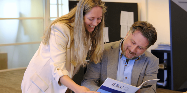 Beau, verpakkingsspecialist bij EBC Golfkarton, aan het werk tijdens een klantbezoek; hij bespreekt een maatwerk doosontwerp met een klant in een magazijn vol verpakkingen.