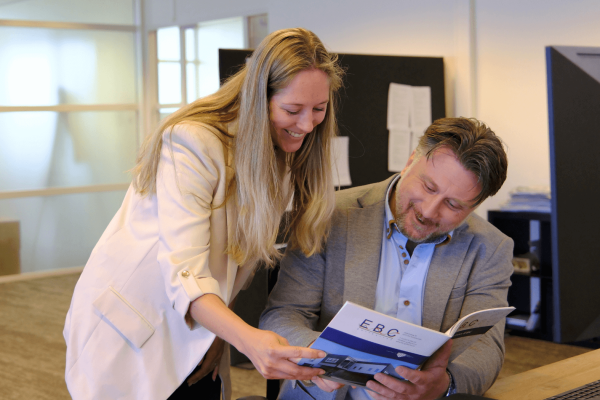 Beau, verpakkingsspecialist bij EBC Golfkarton, aan het werk tijdens een klantbezoek; hij bespreekt een maatwerk doosontwerp met een klant in een magazijn vol verpakkingen.